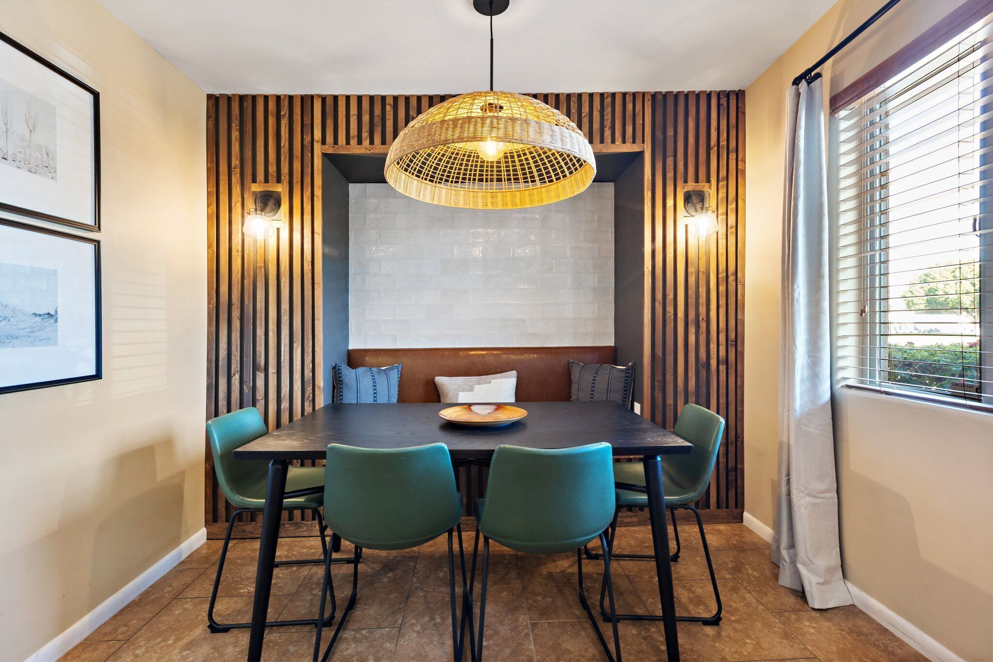 A dining area with a dark wooden table, green chairs, a woven pendant light, and a white brick wall behind the table.