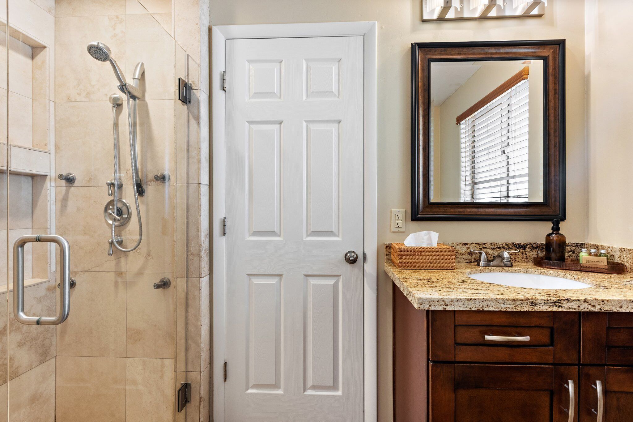 Un baño con una puerta blanca, una ducha con accesorios cromados, un lavamanos con encimera de granito y un espejo encima.