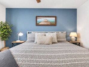Ein Schlafzimmer mit einem Bett, einem Nachttisch, einer Lampe, einem gerahmten Bild an der Wand und einer Pflanze.