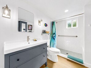 Ein modernes Badezimmer mit einem weißen Waschbecken, einem dunklen Waschtisch, einer Toilette, einer Dusche mit Glastür und einem kleinen Fenster.