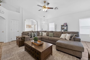 A living room with a sectional sofa, a wooden coffee table, a ceiling fan, and a bookshelf.