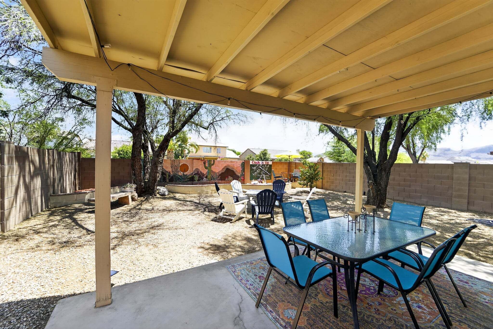 A covered patio with a table and chairs, a concrete floor, and a view of a house and trees in the background.