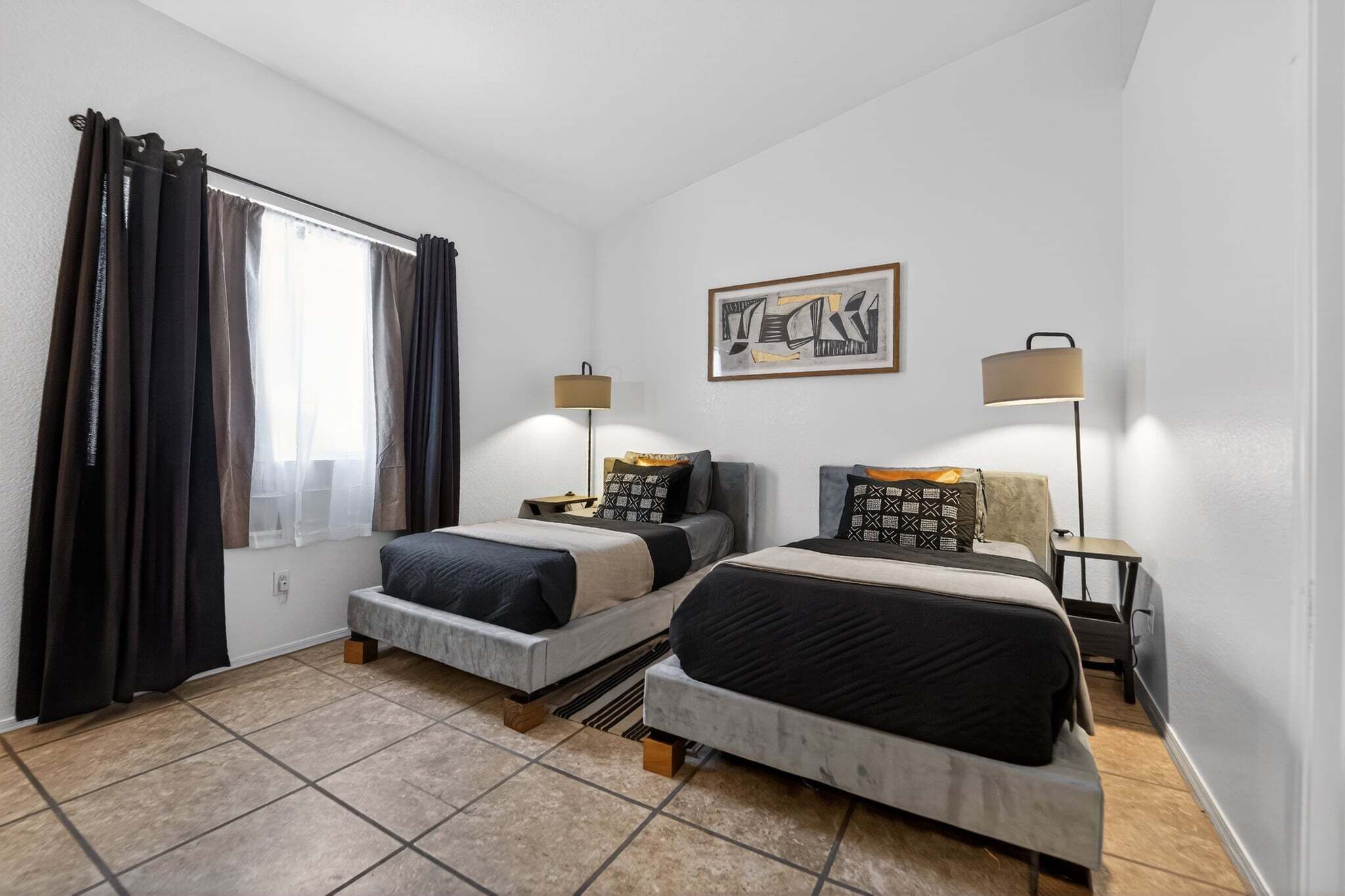 A bedroom with two beds, a window with curtains, a wall-mounted artwork, and bedside lamps.