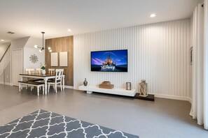 Una sala de estar moderna con un televisor de pantalla plana, una zona de comedor con una mesa de madera y sillas blancas, y una escalera que sube a otro nivel.