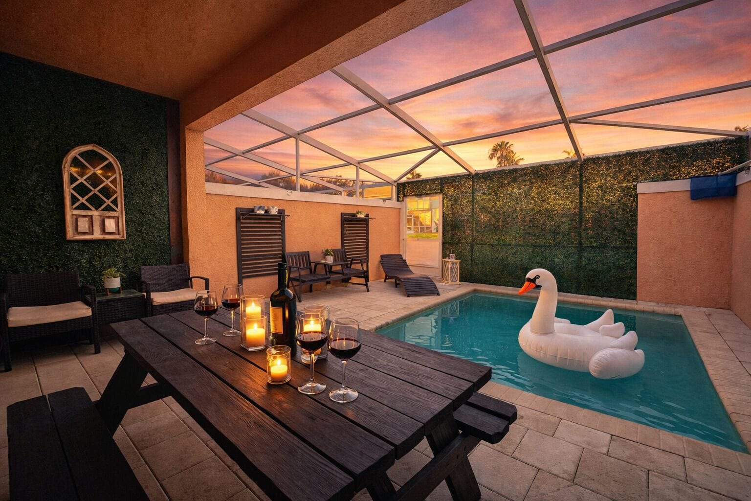 An outdoor pool area with a swan float, a wooden table with glasses and candles, and a view of the sunset.