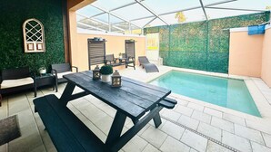 Un área de piscina interior con una mesa y sillas de madera, una piscina pequeña y una pared verde con un ventanal decorativo.