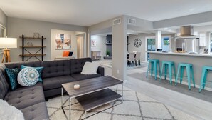 Ein modernes Wohnzimmer mit einem grauen Ecksofa, einem Couchtisch und einem Essbereich, im Hintergrund ist eine Küche zu sehen.