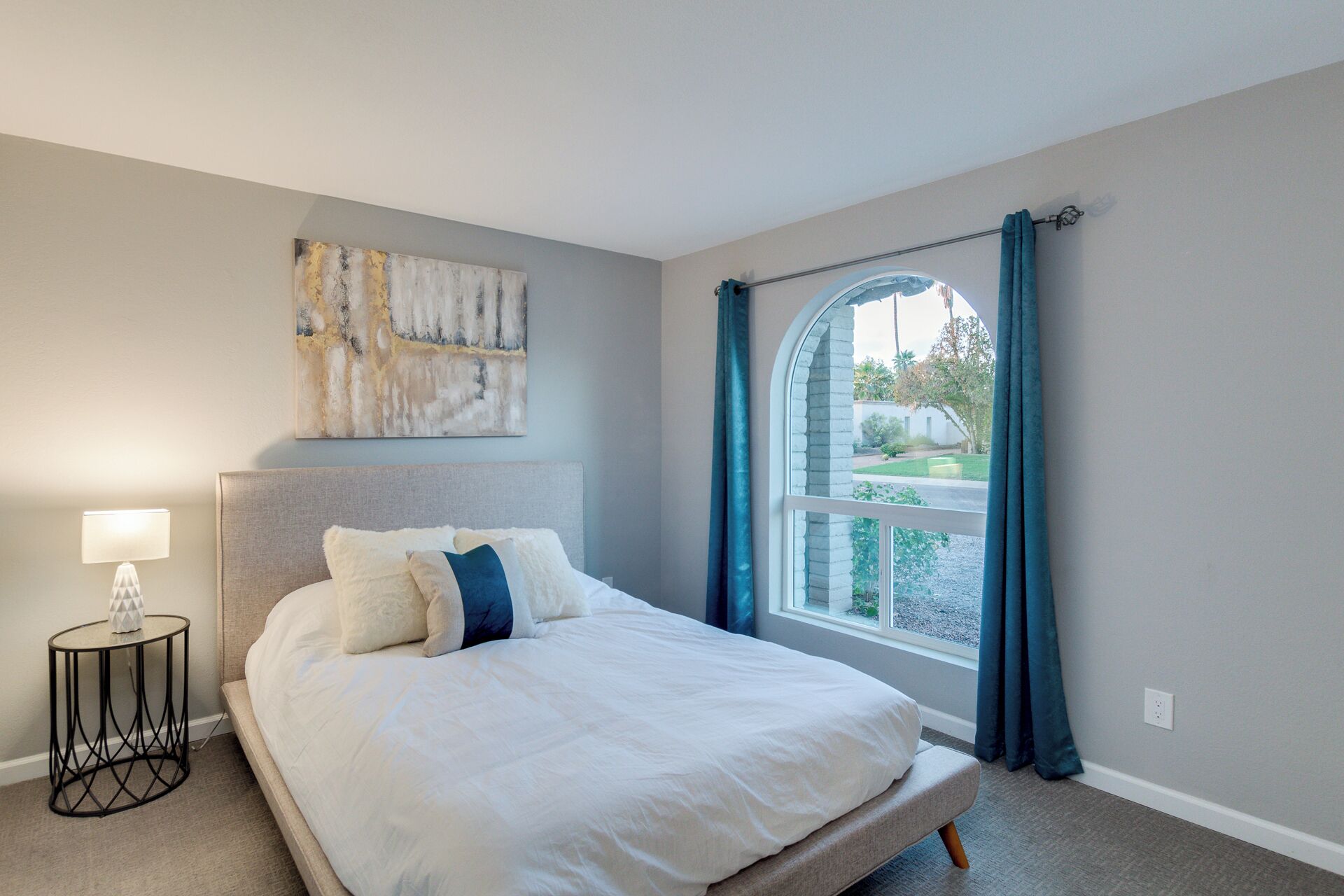 Quarto com cama, mesa de cabeceira, luminária, janela com cortinas e um quadro na parede.