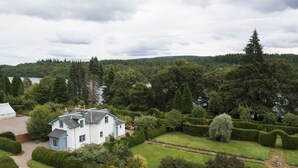 A large house surrounded by a well-maintained garden with a pond, a greenhouse, and a view of a lake and forested hills.