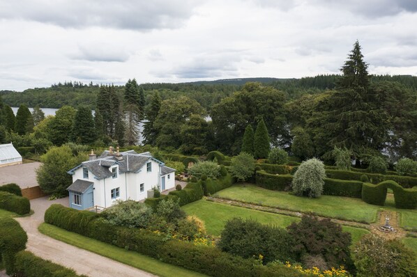 Una casa grande rodeada de un jardín cuidado con un estanque, un invernadero y vistas a un lago y colinas boscosas.