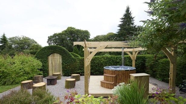 A wooden pergola with a hot tub, wooden benches, and a wooden deck in a garden setting.