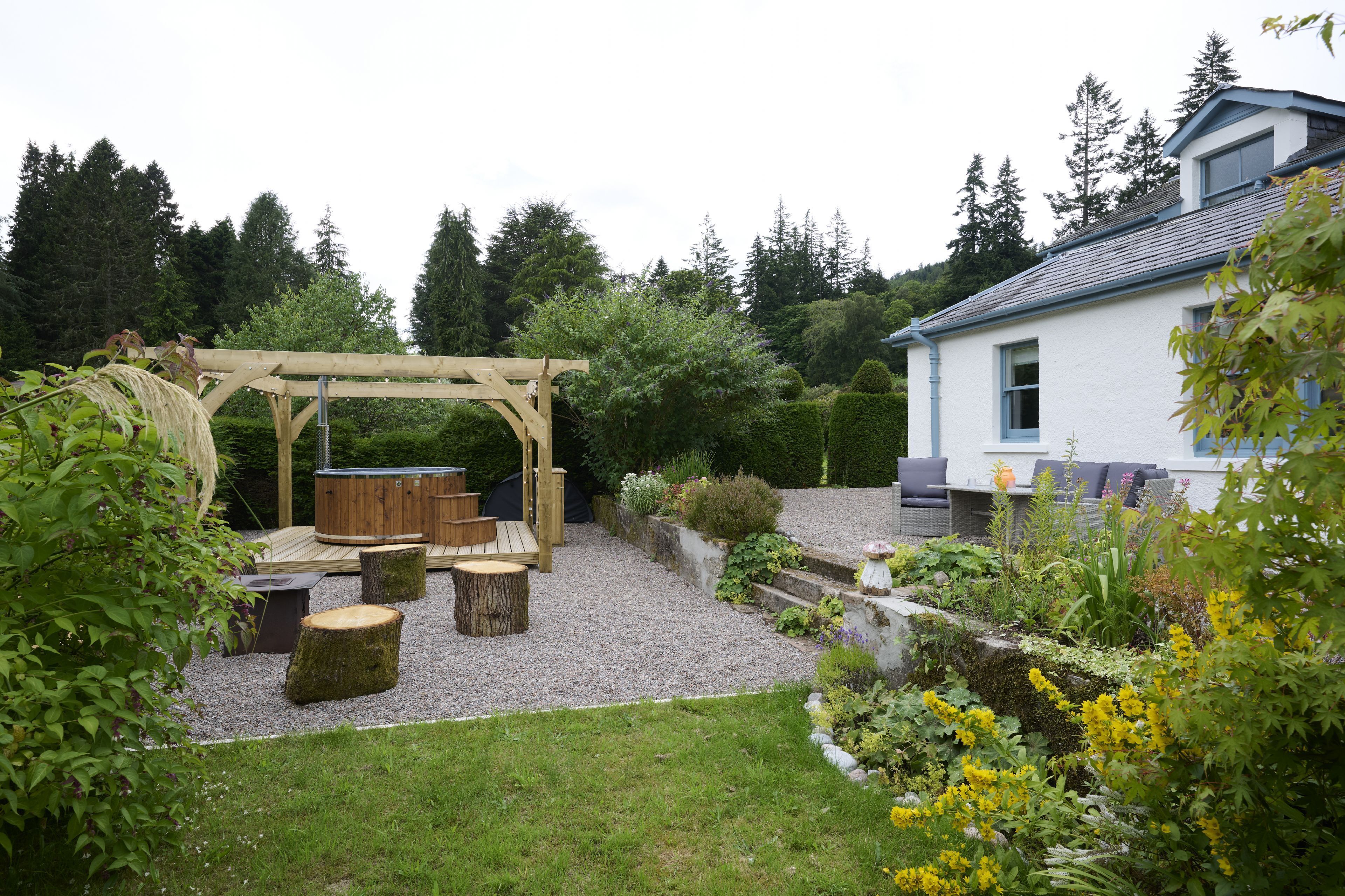 Une pergola en bois avec des sièges, une terrasse en gravier et un jardin agrémenté de diverses plantes et fleurs.