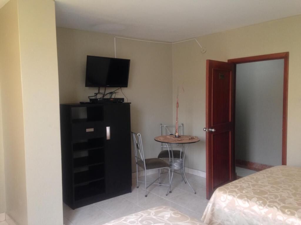 Una habitación pequeña con cama, televisión sobre un mueble bajo, una mesita con una silla y una puerta.