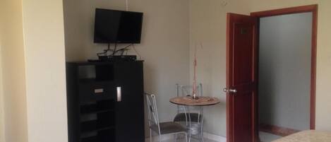 Una habitación pequeña con cama, televisión sobre un mueble bajo, una mesita con una silla y una puerta.