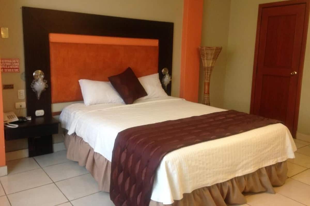 A neatly made bed with a headboard, a bedside table with a lamp, and a door in the background.