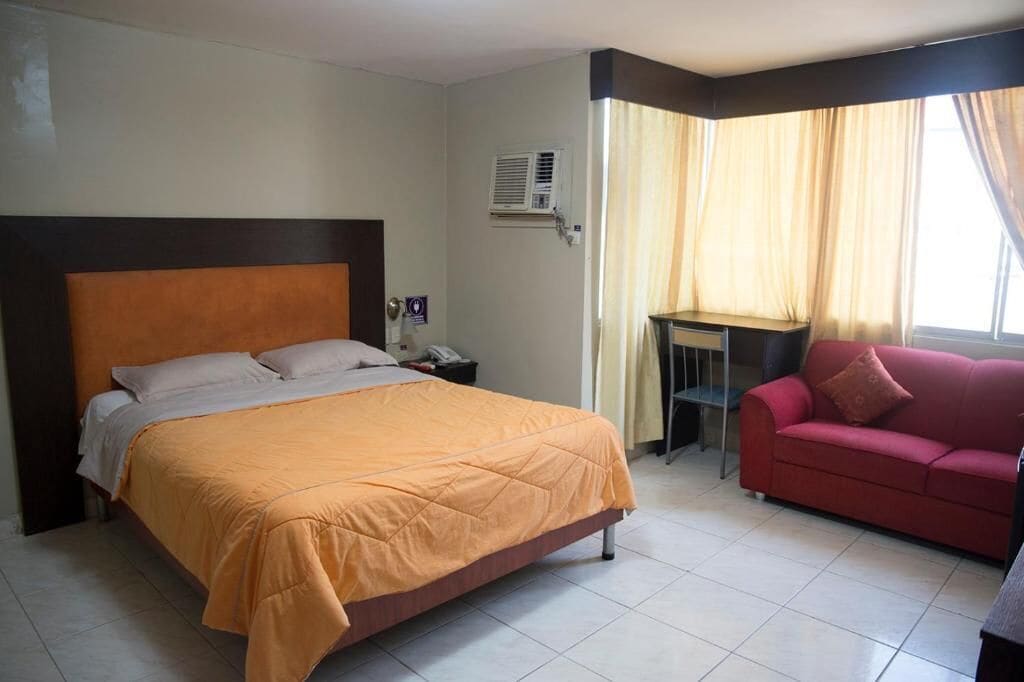 A hotel room with a bed, a desk, a red sofa, and a window with curtains.