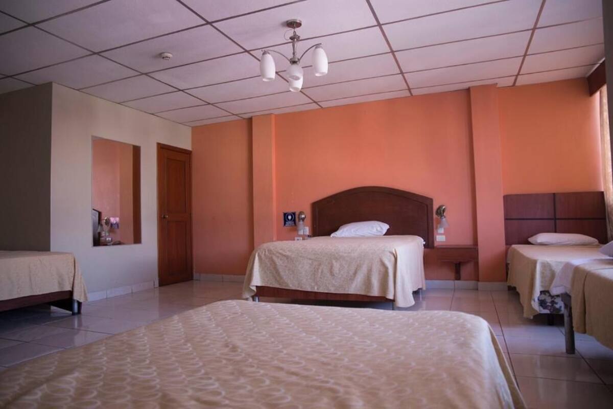 A hotel room with a large bed, two smaller beds, a wooden headboard, a nightstand, and a ceiling light fixture.
