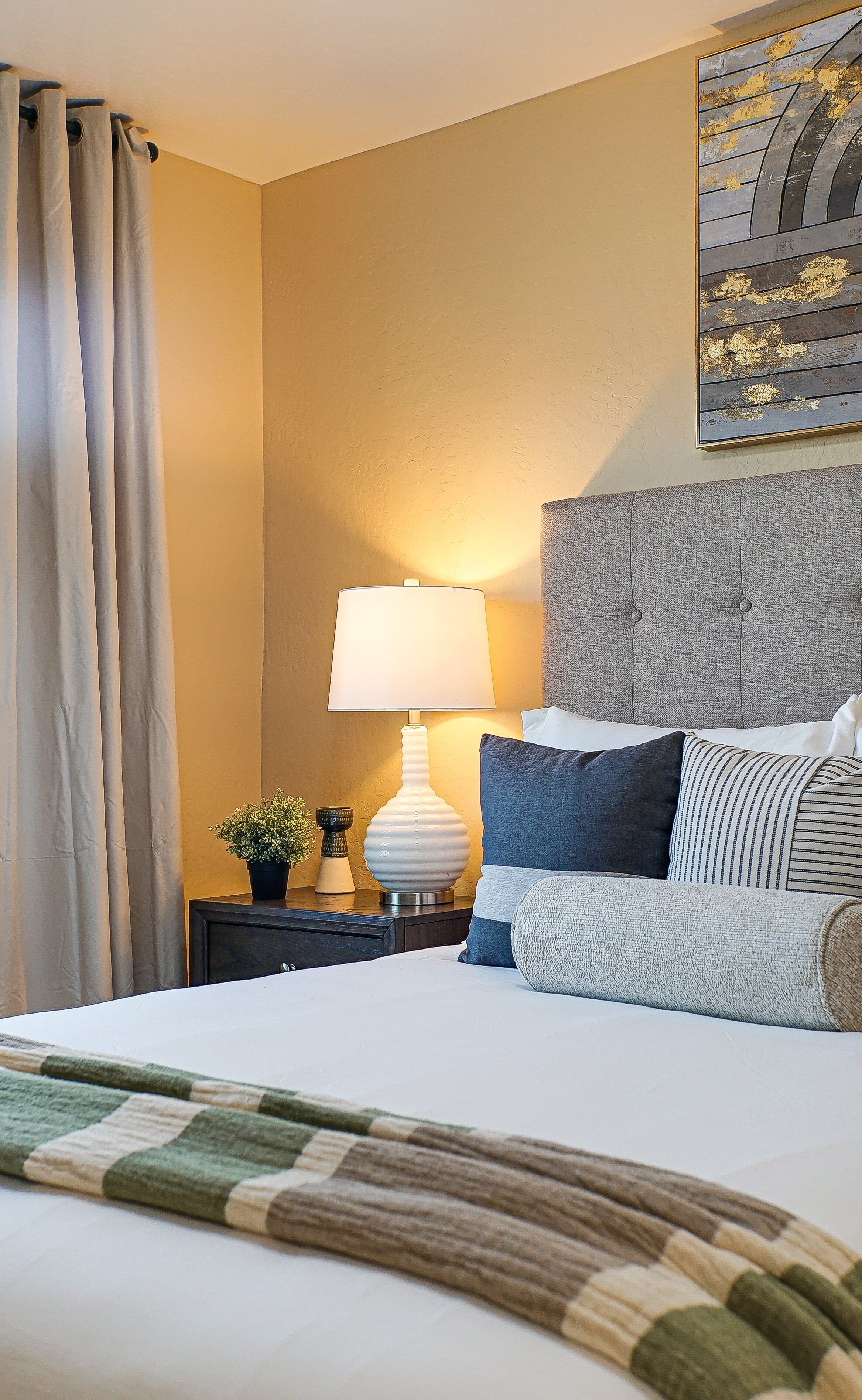A hotel room with a bed, a nightstand, a lamp, a potted plant, and a painting on the wall.