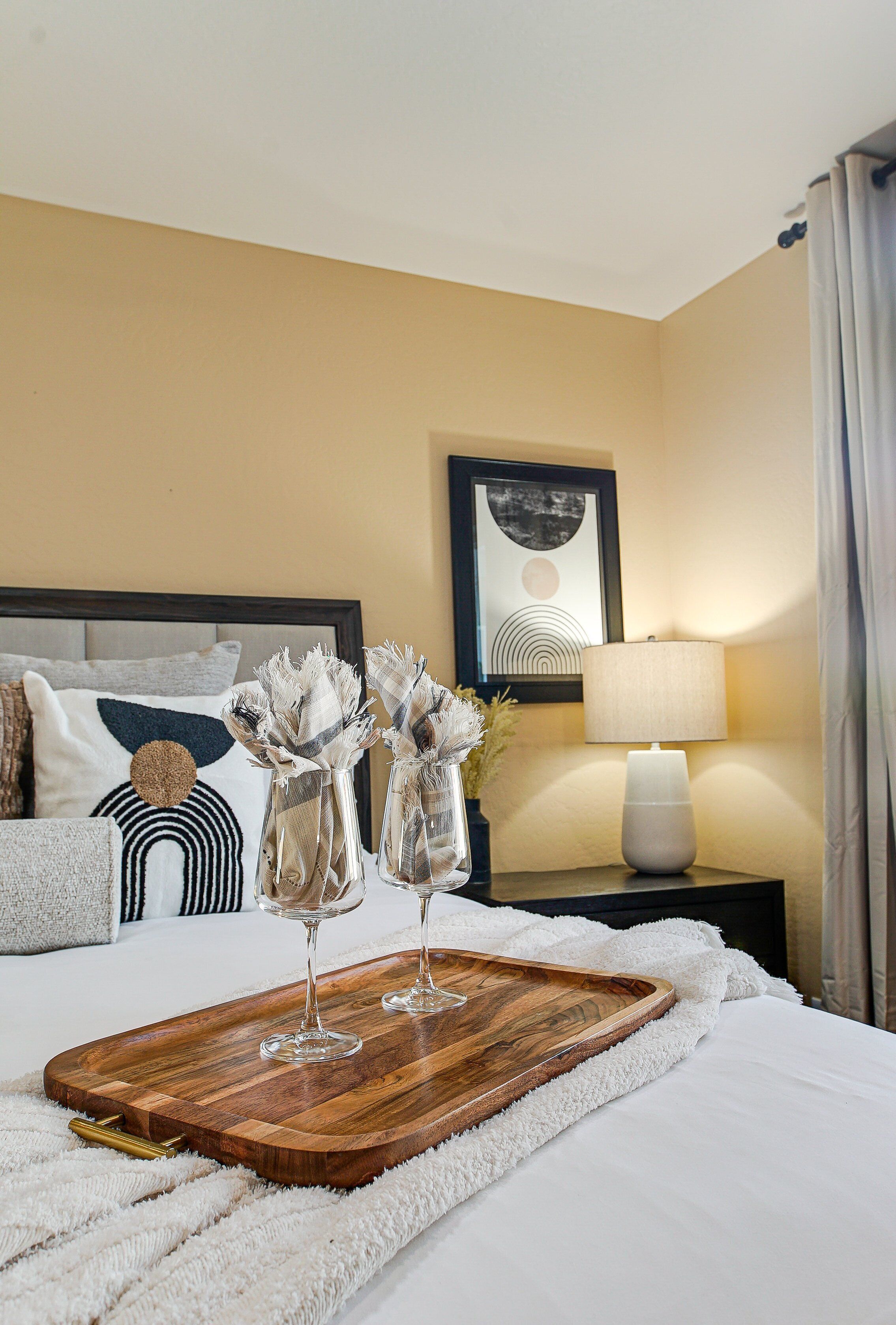 Una cama bien hecha con una bandeja de madera, dos jarrones de vidrio con flores secas, una lámpara y un cuadro en la pared.