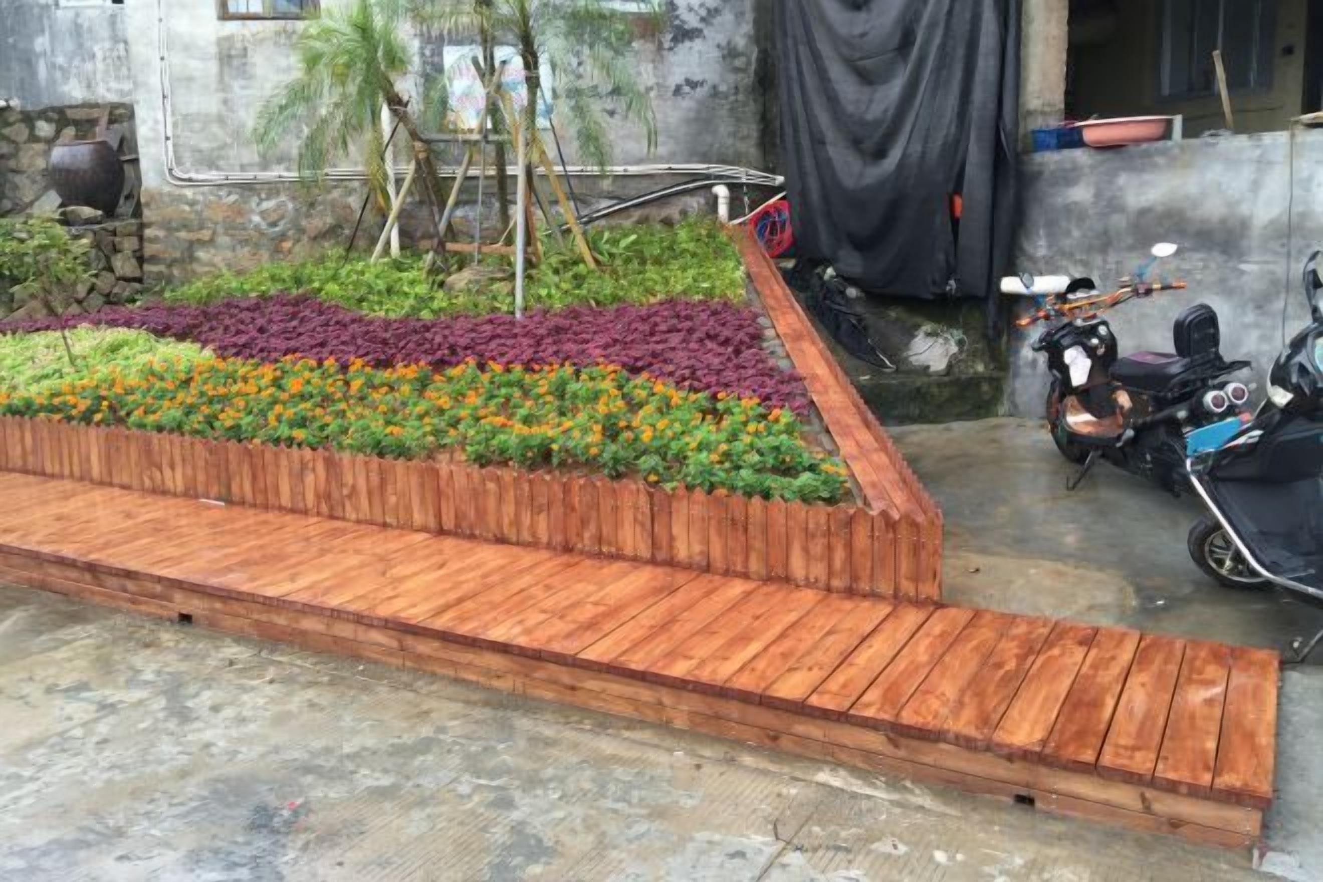 Una terraza de madera con un jardín de flores, una motocicleta y una superficie de hormigón.