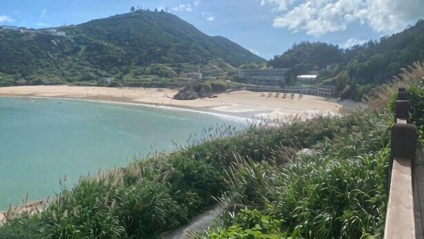 Paisagem costeira com uma praia, uma montanha e um edifício ao longe.