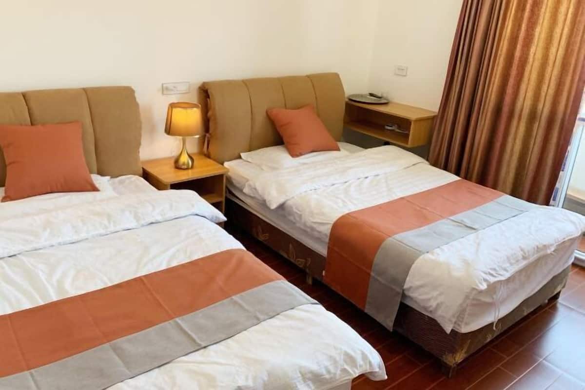 A hotel room with two beds, a nightstand with a lamp, a wooden side table, and a window with curtains.