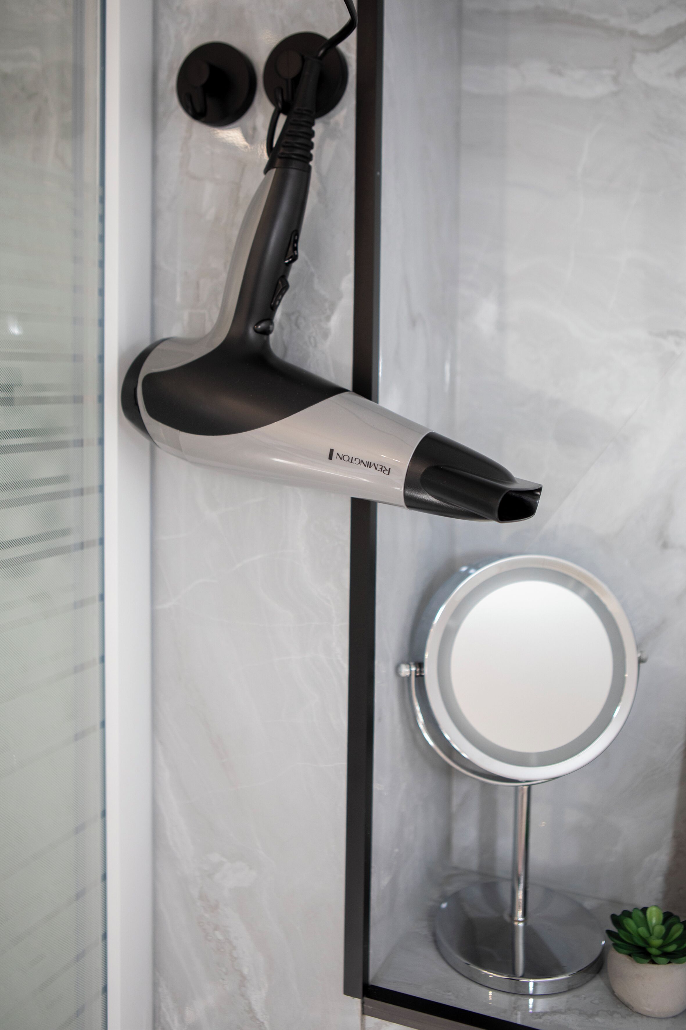 Une salle de bain avec un sèche-cheveux fixé au mur, un miroir rond et une petite plante en pot.