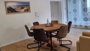 Een moderne eetruimte met een houten tafel en stoelen, een schilderij van een strand en uitzicht op de natuur door een glazen deur.