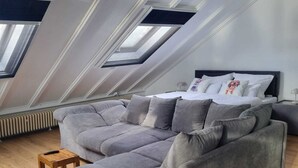 Een moderne loft slaapkamer met een grote grijze modulaire bank, een klein houten krukje en een bed met een donker hoofdbord. De kamer heeft een schuine kap met dakramen en een radiator onder het raam.