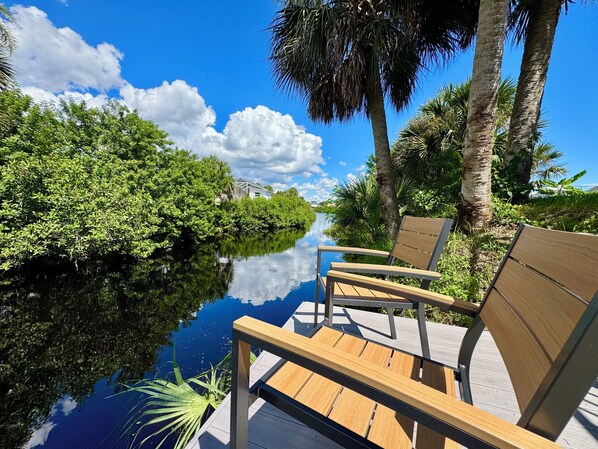 A wooden deck with a bench overlooking a calm water body, surrounded by lush greenery and palm trees.