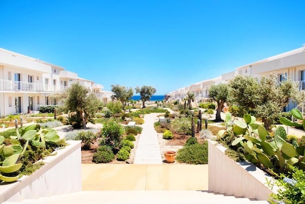 A resort with a central pathway leading to a beach, surrounded by white buildings and various plants.