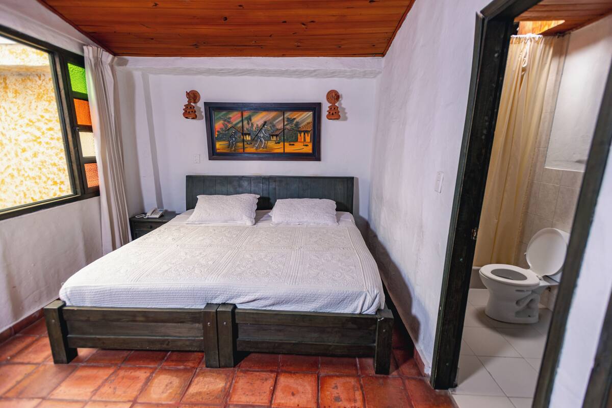 A bedroom with a wooden bed, a painting on the wall, a window with curtains, and a toilet visible through an open door.