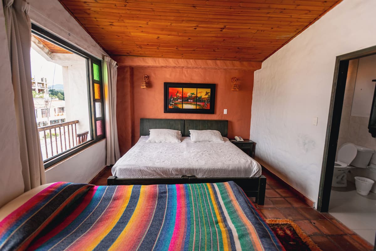 A bedroom with a bed, a window with curtains, a nightstand, a painting on the wall, and a view of the outside.