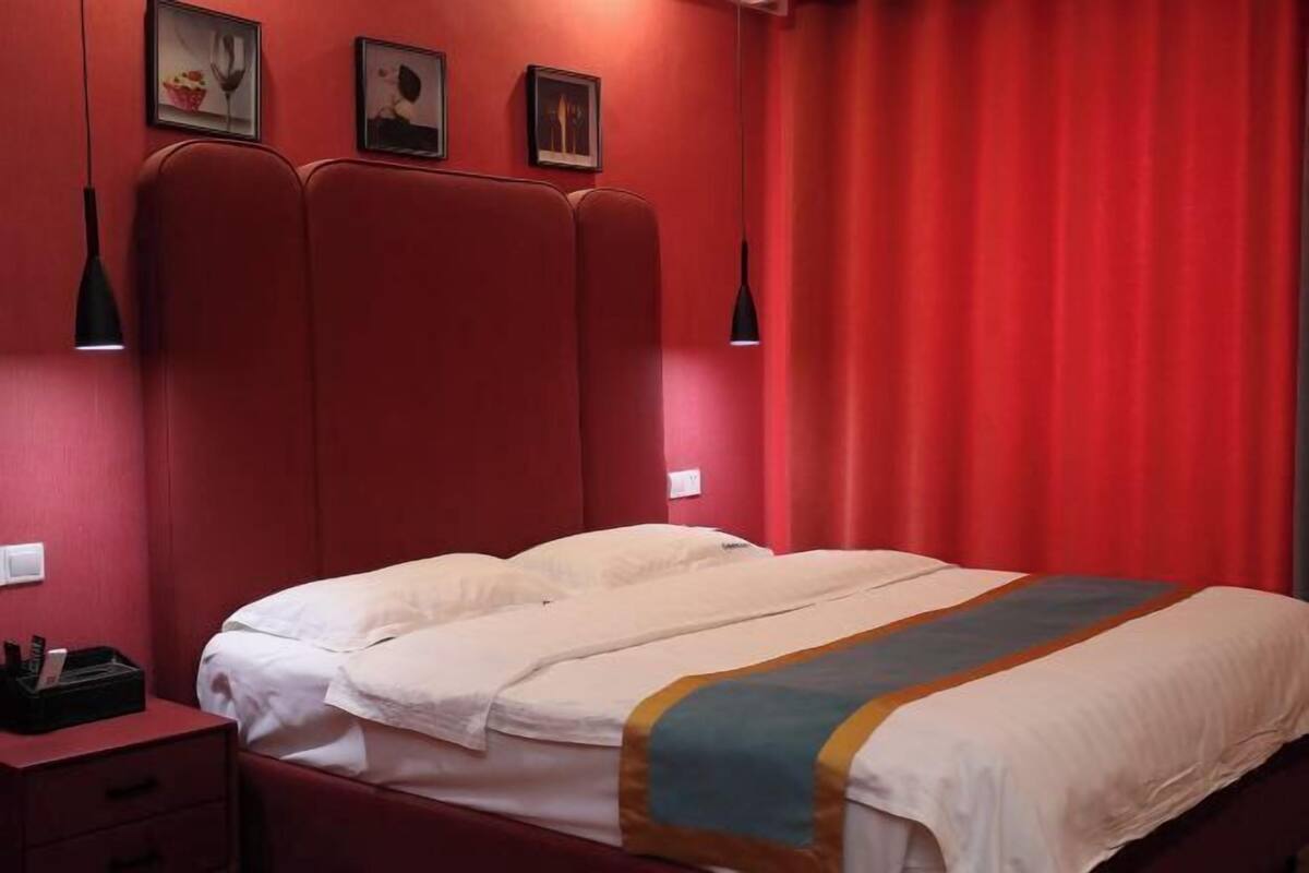 Habitación de hotel con pared roja, cama con ropa blanca y una manta multicolora, mesita de noche con lámpara, y tres cuadros en la pared.