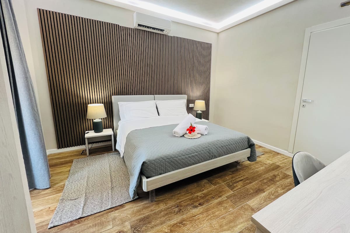 A modern hotel room with a bed, bedside lamps, a desk, and a wooden floor.