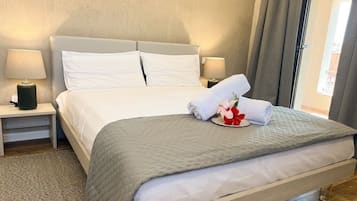 A neatly made bed with a grey quilt, white pillows, and a floral arrangement on the bed. There are two bedside tables with lamps, and a window with curtains.