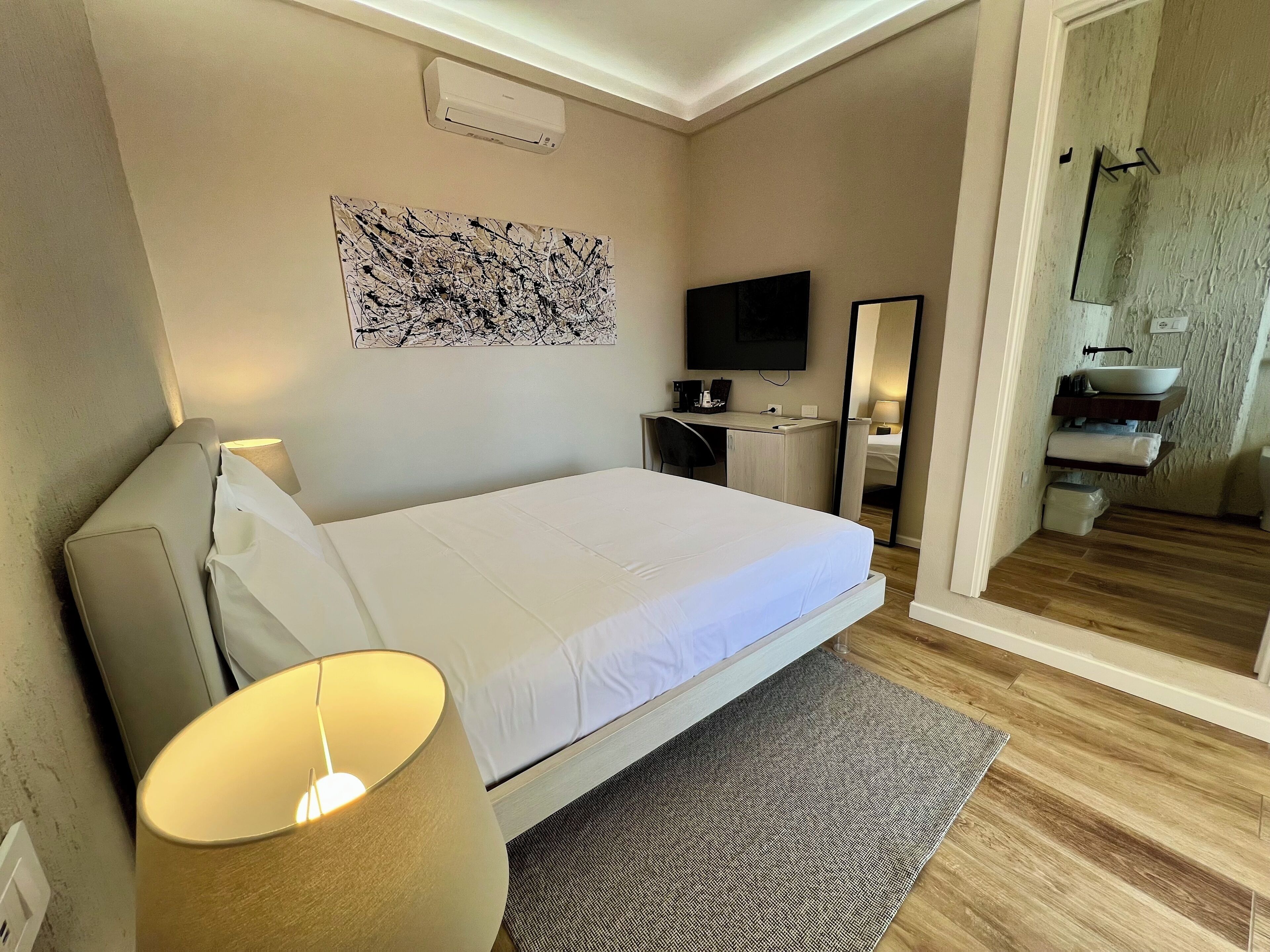 A modern hotel room with a bed, bedside lamp, desk, and a flat-screen TV.