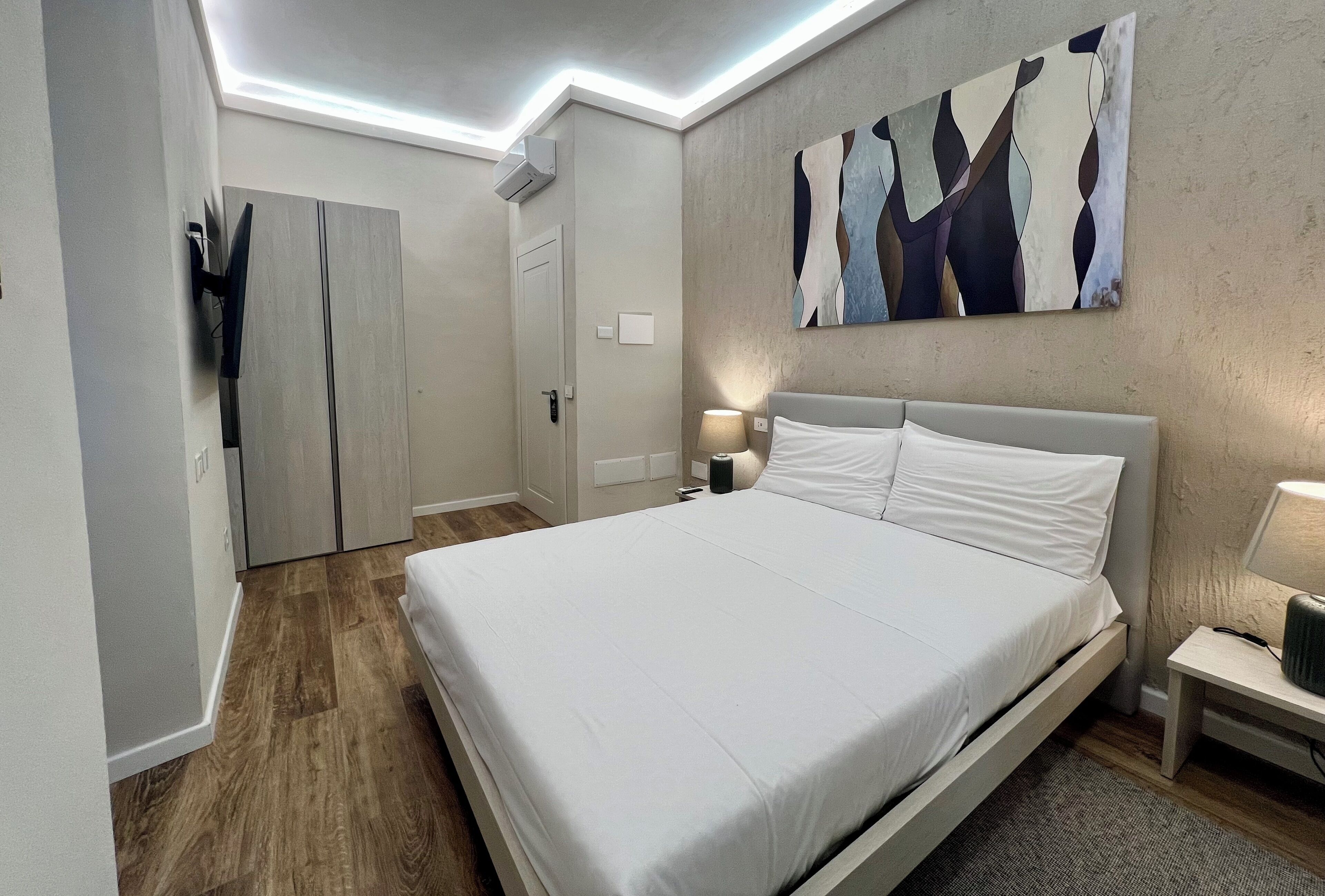 Een moderne hotelkamer met een groot bed, een kledingkast, een schilderij aan de muur en een nachtkastje met een lamp.