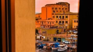 A view from a window overlooking a harbor with boats, buildings, and a pink sky. - Maison Madeleine (La Maddalena)