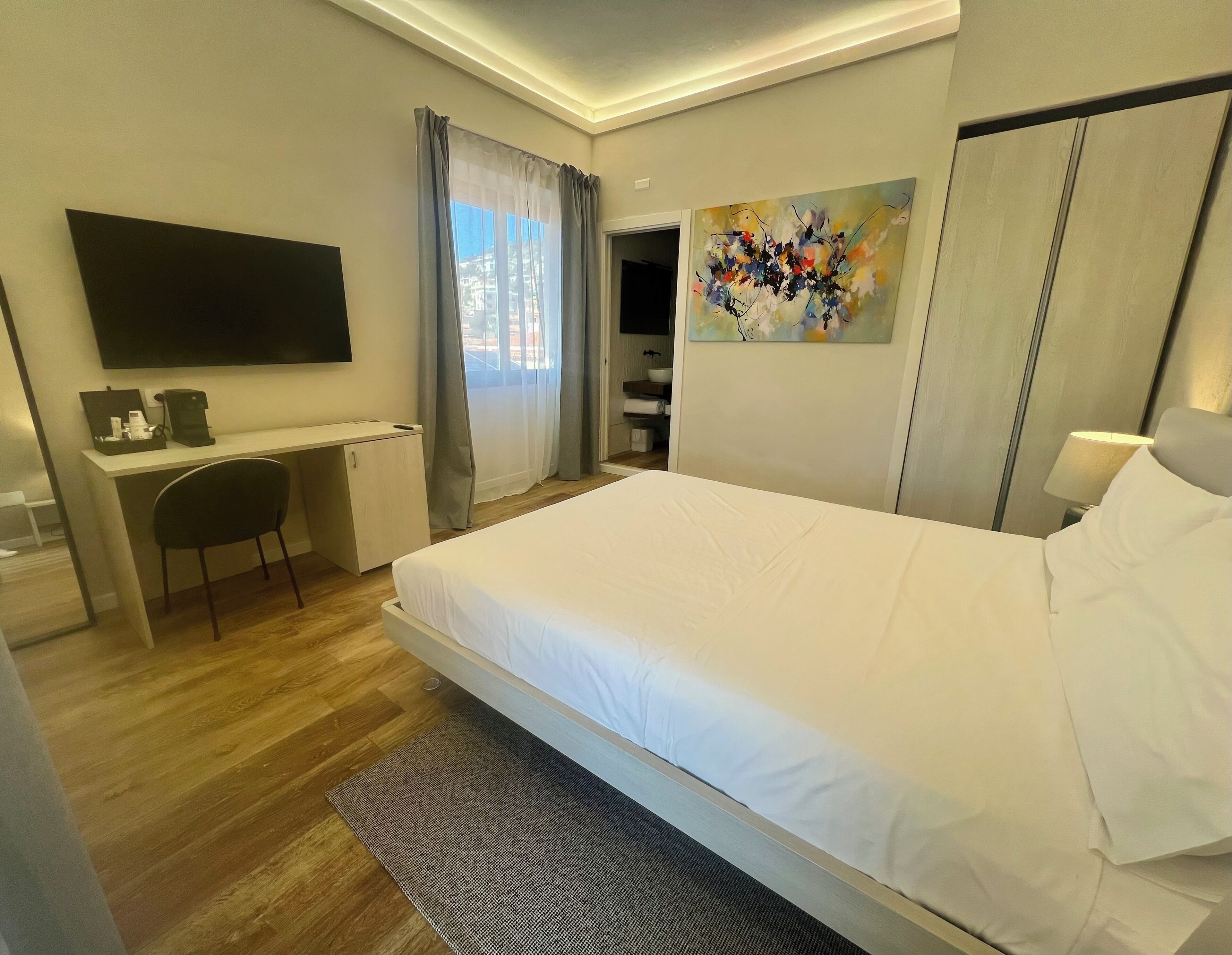 A hotel room with a bed, desk, chair, TV, artwork, and a bathroom visible through an open door.