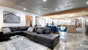 Een moderne woonkamer met een hoekbank, een glazen salontafel en een eetgedeelte met blauwe stoelen.