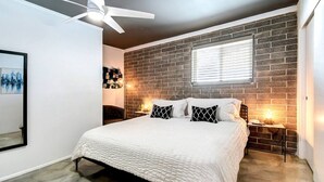 Een slaapkamer met een bakstenen muur, een bed met witte beddengoed, een plafondventilator en een raam met zonwering.