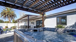 Een hottub onder een houten pergola, een terras met stoelen, en een huis op de achtergrond.