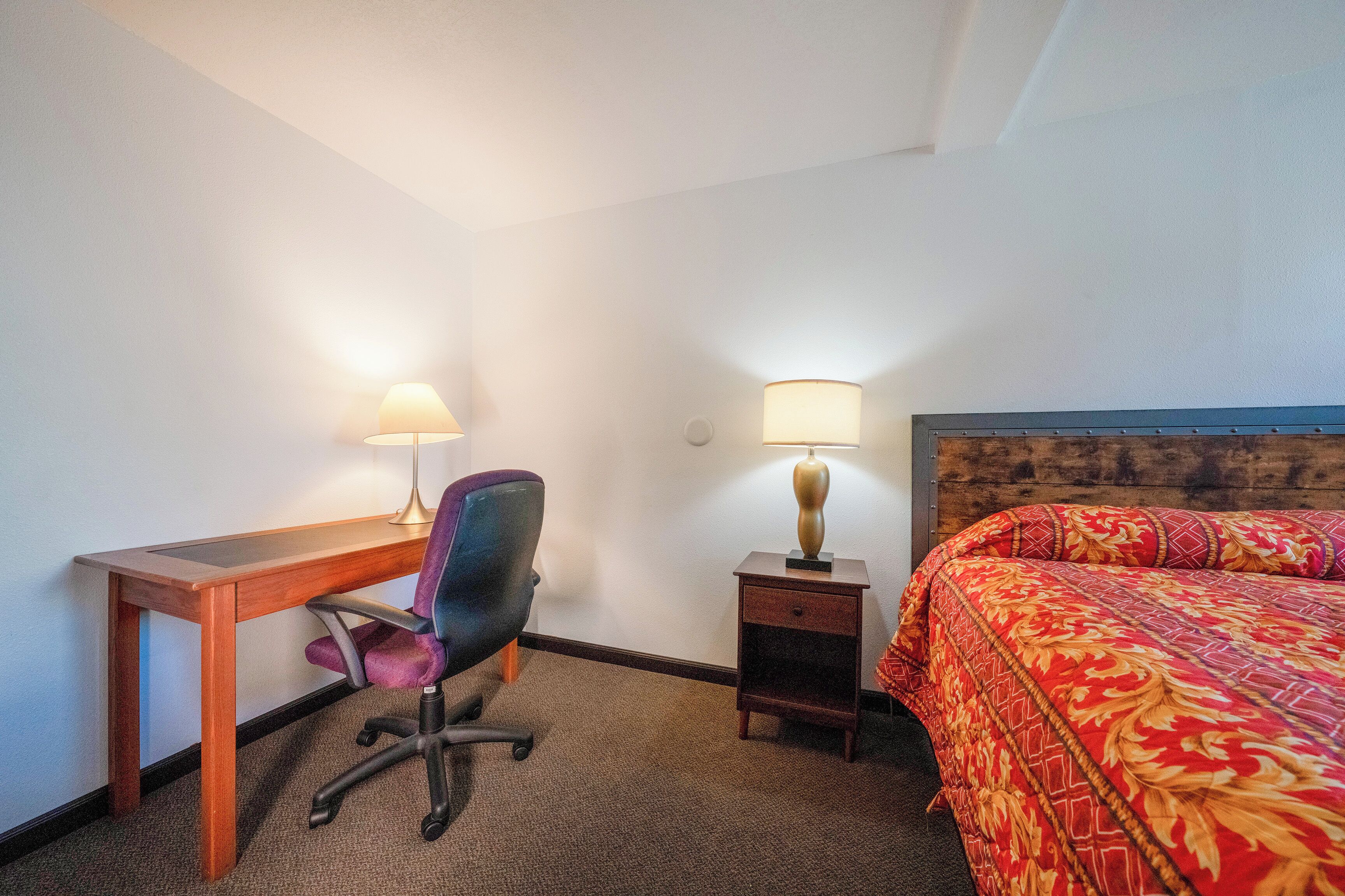 A hotel room with a wooden desk, a purple office chair, a small bedside table with a lamp, and a bed with a red and gold patterned bedspread.