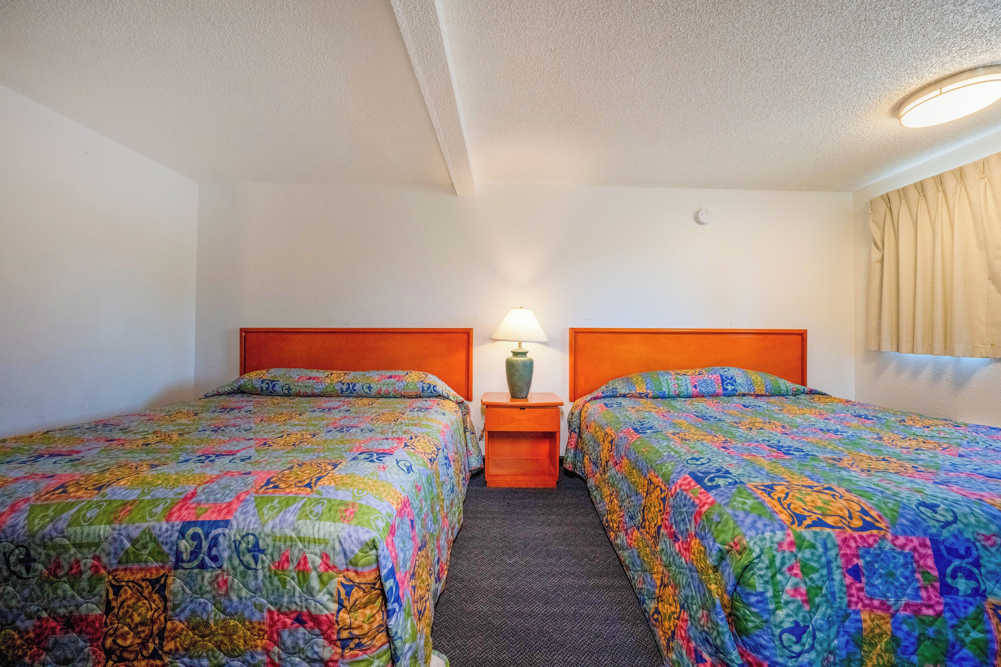 A hotel room with two beds, each with colorful quilts, a small table with a lamp, and a window with curtains.
