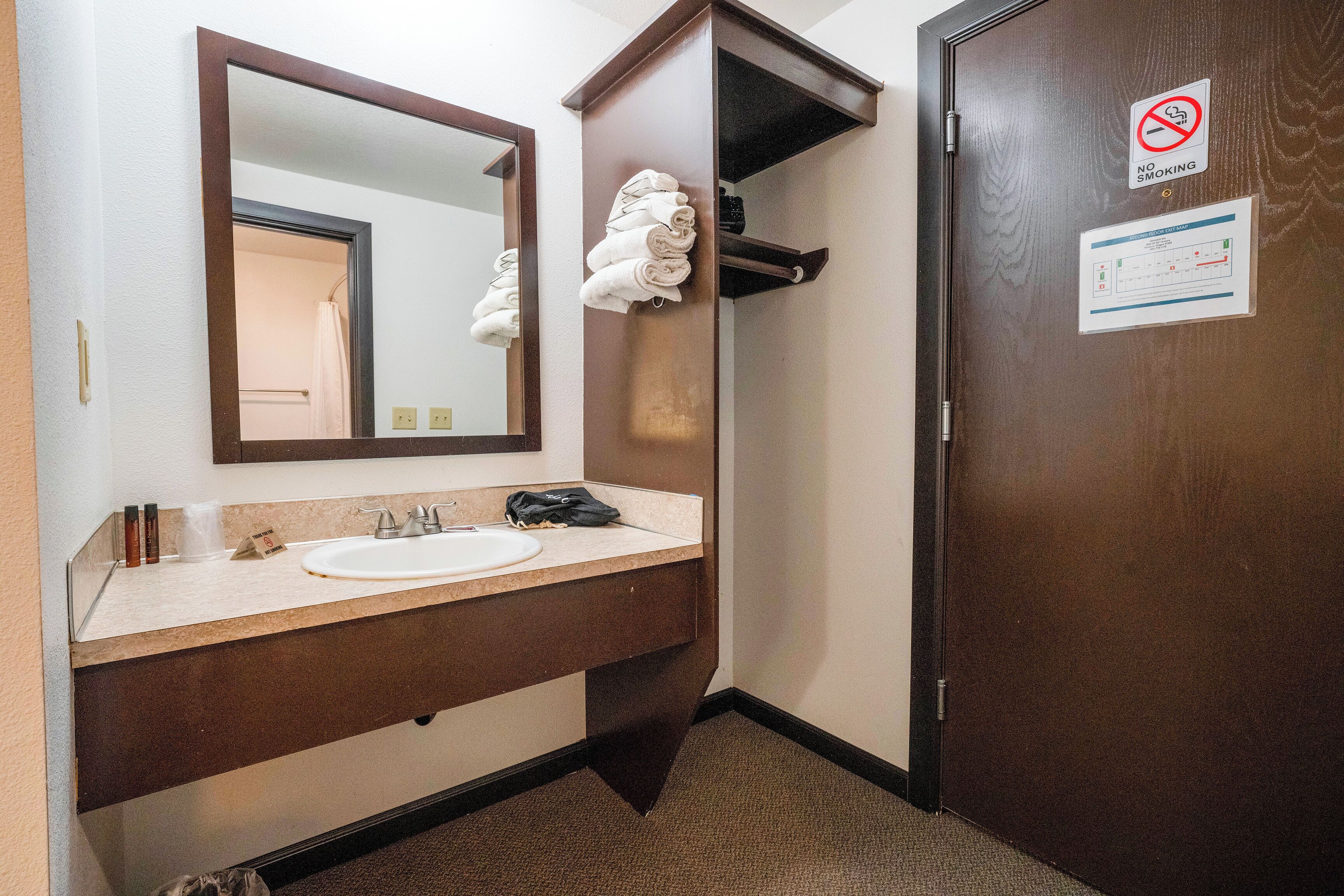A bathroom with a sink, mirror, towel rack, and a door with a no-smoking sign.