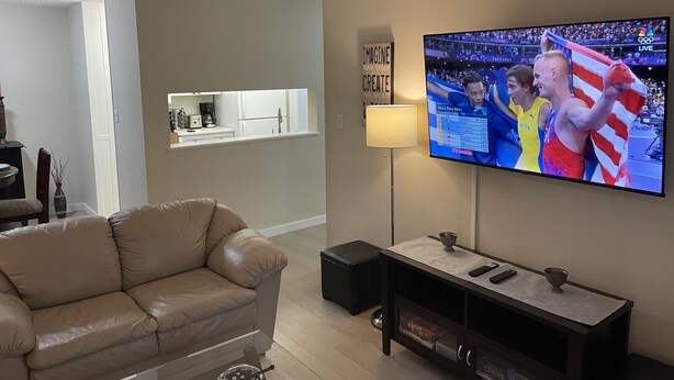Una sala de estar con un sofá color beige, un televisor en la pared mostrando un evento deportivo, una mesita de noche con una lámpara, y una cocina al fondo.