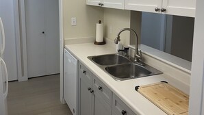 Una cocina con armarios blancos, un lavamanos de acero inoxidable, una tabla de cortar de madera y un techo blanco.