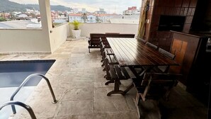 Una terraza en la azotea con una mesa de madera larga y bancos, una piscina pequeña y un televisor instalado en una pared de ladrillo.
