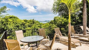 A terrace with a round table and chairs, surrounded by a metal railing, overlooking a lush landscape and a distant view of the sea. - Ocean View 2Br/2Bth Condo In Tamarindo (Tamarindo)
